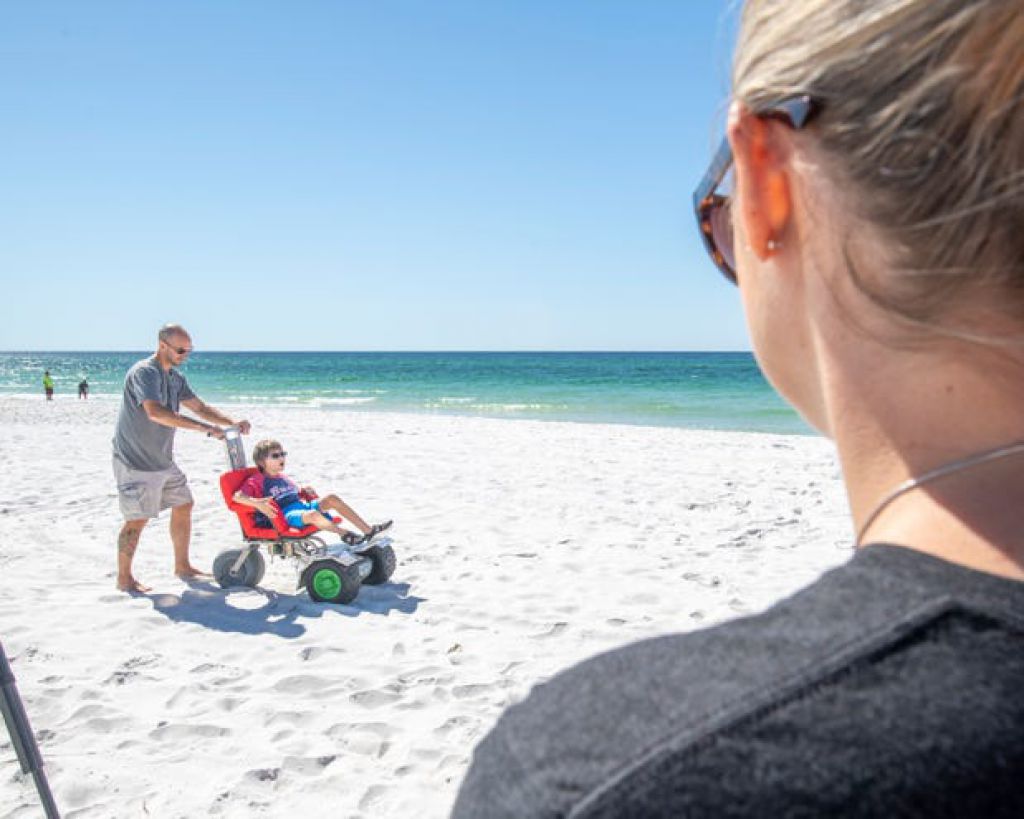 朱莉·丹顿（Julie Denton）看著她的丈夫特雷弗·丹顿（Trevor Denton）在纳瓦拉海滩的沙滩上沿著特制的电动轮椅推著他们11岁的儿子克里斯蒂安（Christian）。