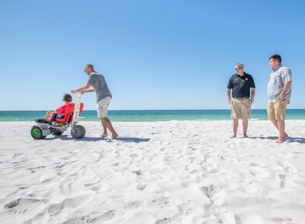 照片人Shane Kahl（右）和Navarre Beach Fire Rescue首席Danny Fureigh观看时，特雷弗·丹在Navarre Beach的特制电动轮椅上推著他11岁的儿子Chr
